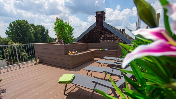A wooden deck with outdoor seating, a brick fireplace, and a garden area with plants and flowers. - Hotel Dampfmühle (Neukirchen-Vluyn)