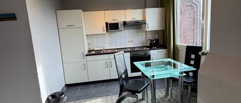Una cocina compacta con armarios blancos, un microondas y una estufa a gas. Hay una mesa de comedor con superficie de vidrio y dos sillas.