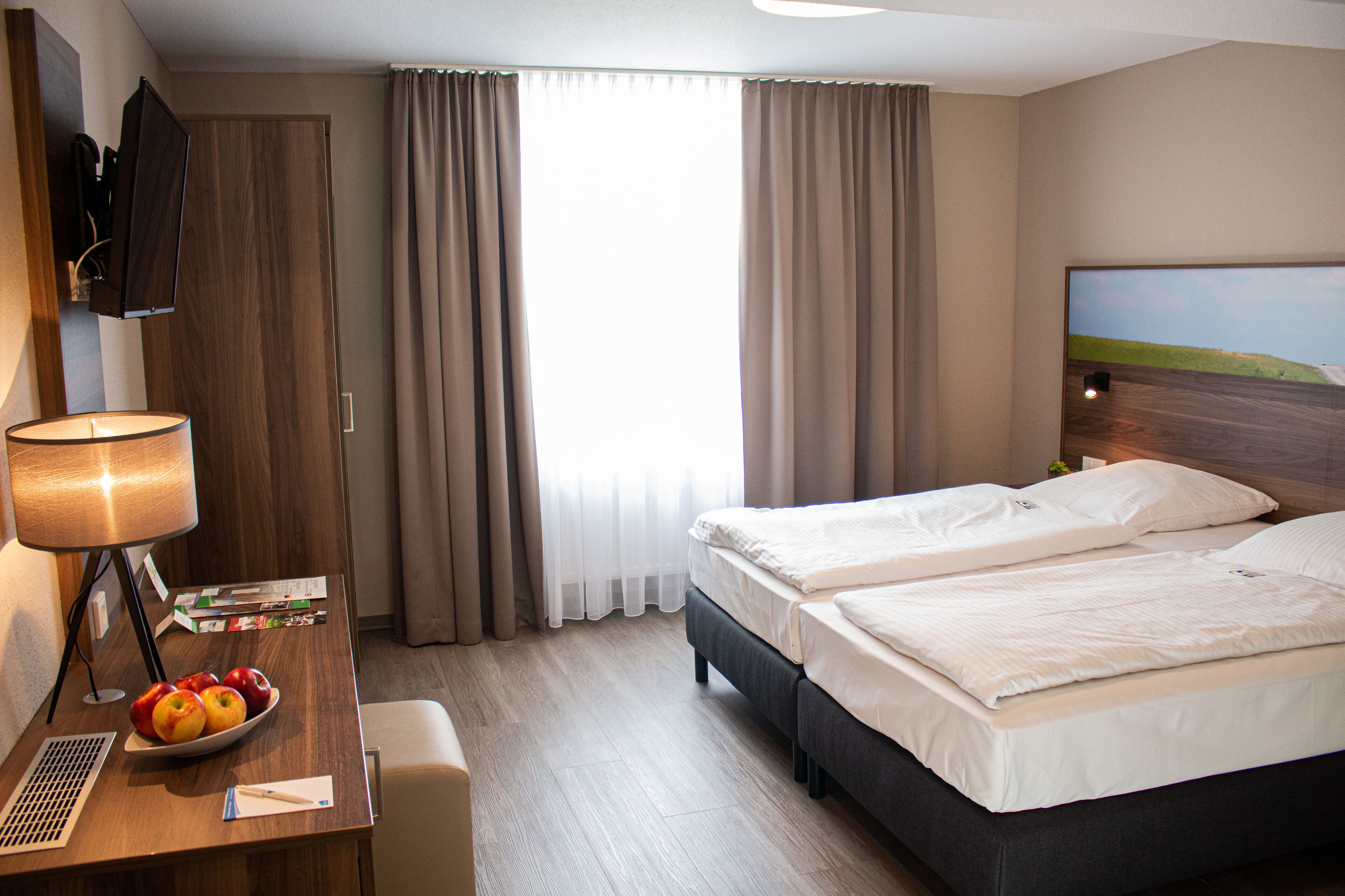 Een hotelkamer met twee bedden, een bureau met lamp, een schaal fruit en een televisie aan de muur.