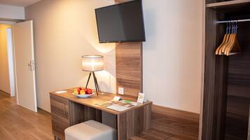 A hotel room with a wooden desk, a flat-screen TV mounted on the wall, a lamp, a bowl of fruit, and a wooden coat rack.