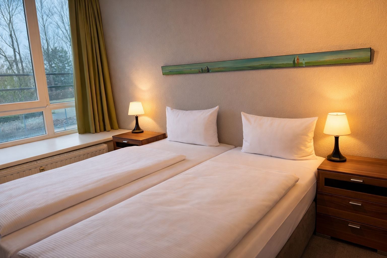 Ein Hotelzimmer mit einem Bett, zwei Nachttischen, einer Lampe auf jedem Tisch, einem Fenster mit Vorhängen und einem Gemälde an der Wand.