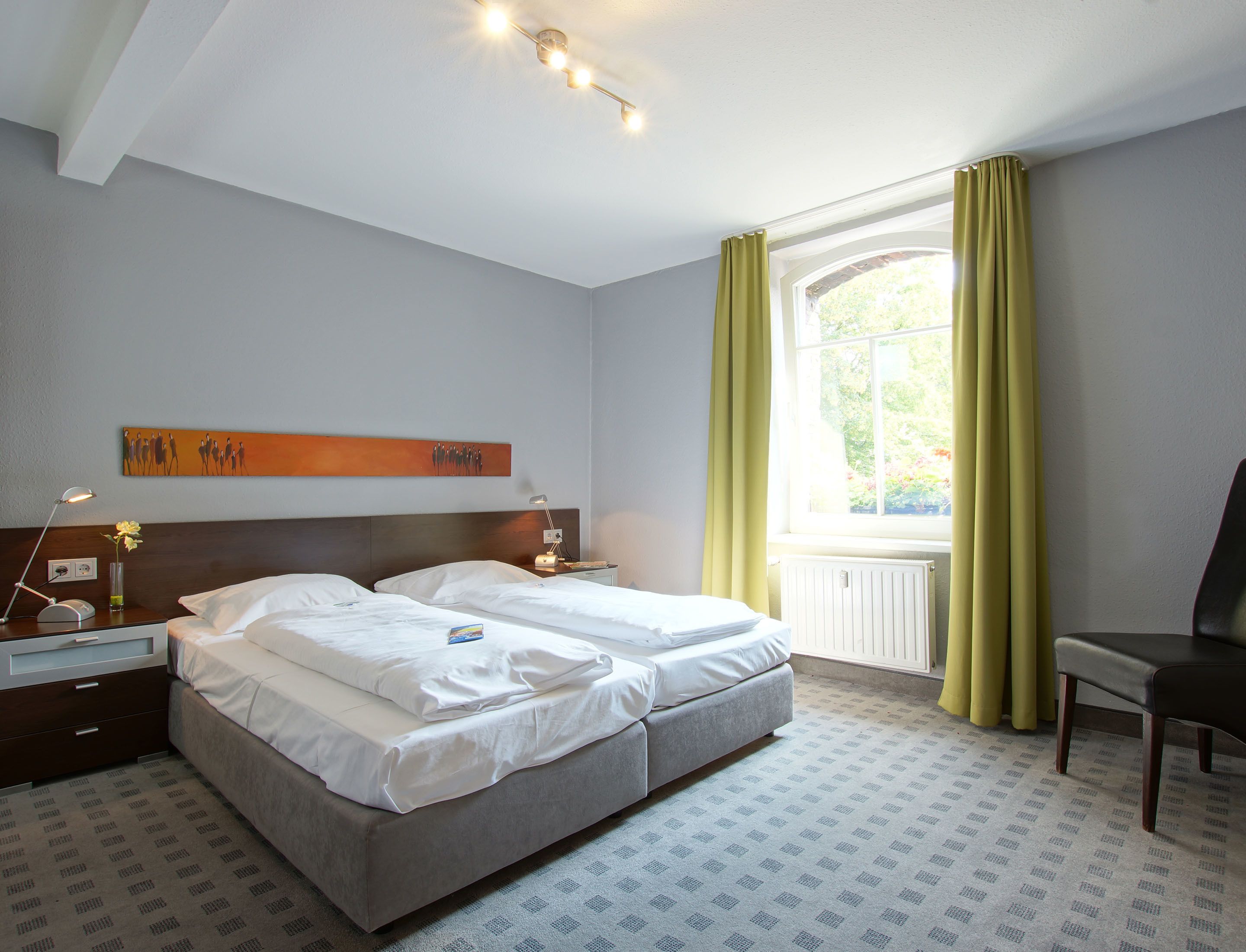 A hotel room with a double bed, two bedside tables with lamps, a window with curtains, and a chair.