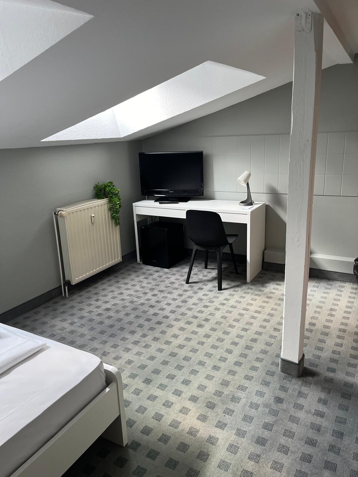 Ein schlichtes Zimmer im Studentenwohnheimstil mit Dachfenster, Schreibtisch mit Computer, Stuhl, Heizkörper und Bett.