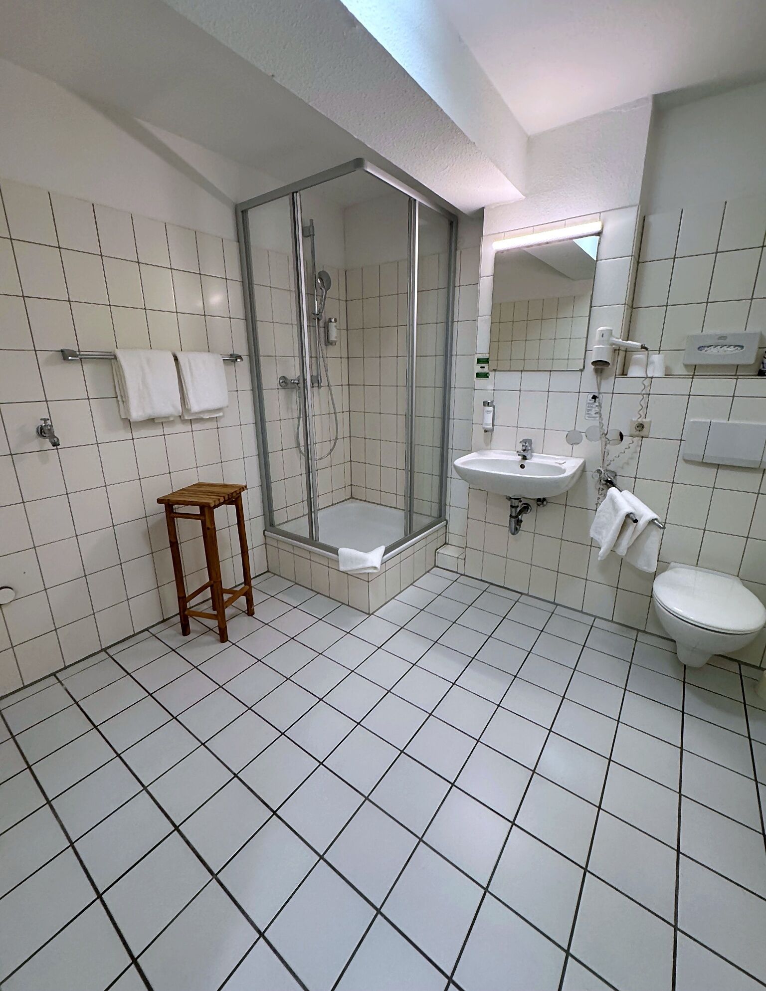 A bathroom with a shower, sink, toilet, and a wooden stool.
