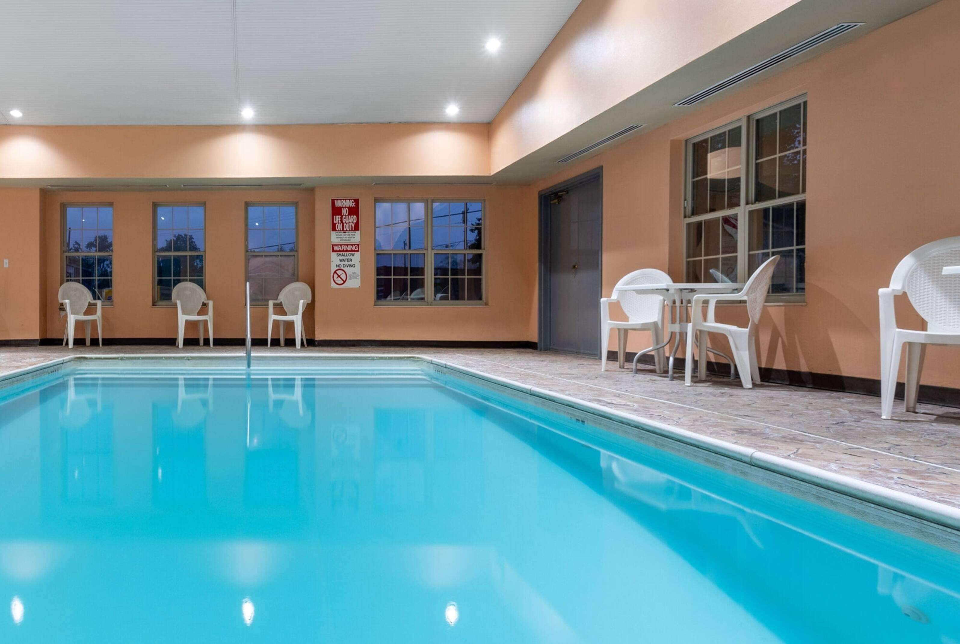 An indoor swimming pool with clear blue water, surrounded by white chairs and tables, and large windows with a view of the outside.