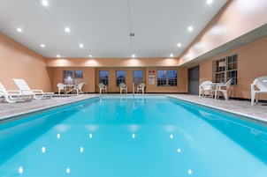 Piscina con sillas de descanso y mesas, rodeada de muros y ventanas color beige.