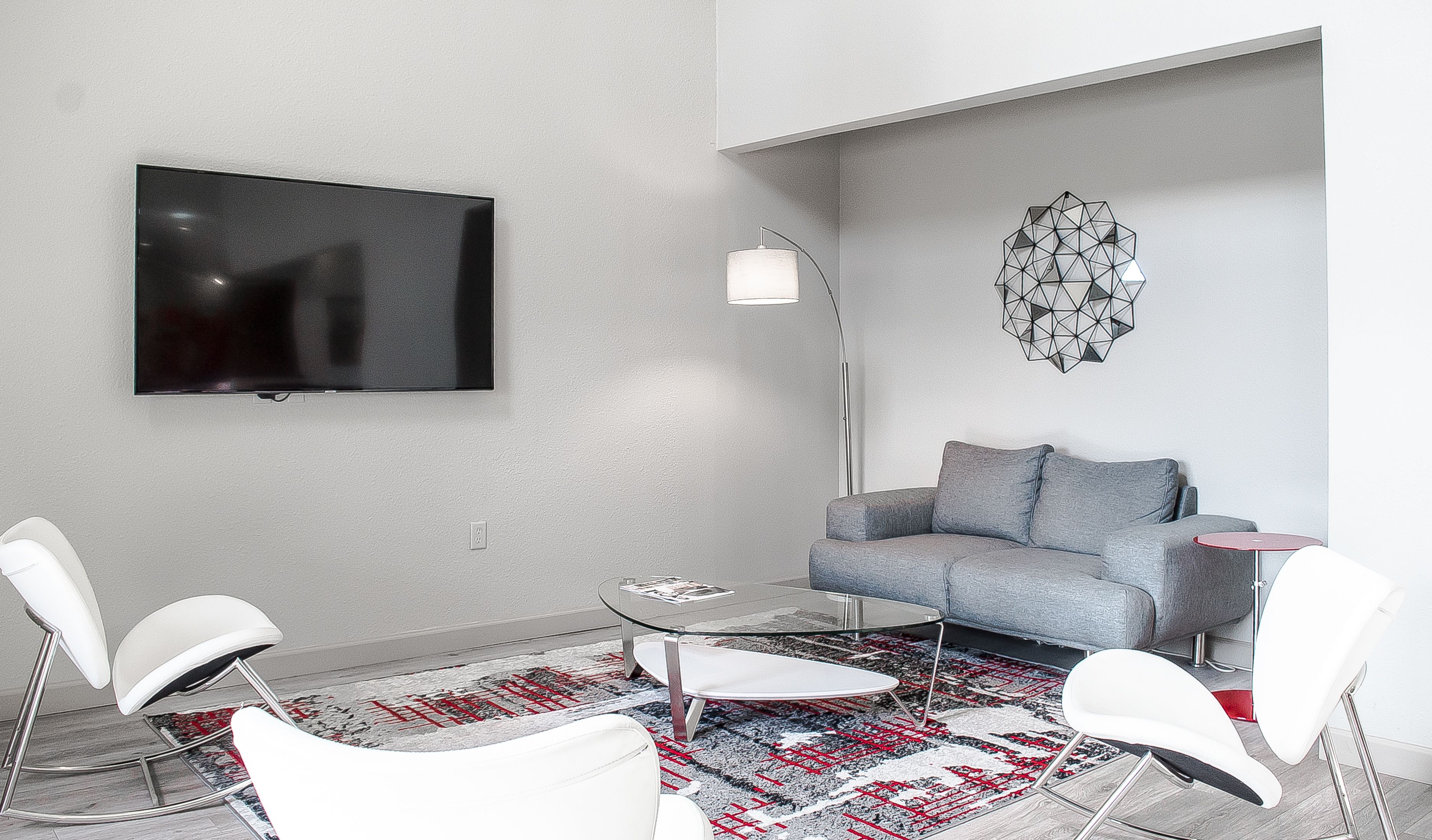 Una sala de estar moderna con un sofá gris, un televisor de pantalla plana montado en la pared, una lámpara de pie y un adorno mural geométrico.
