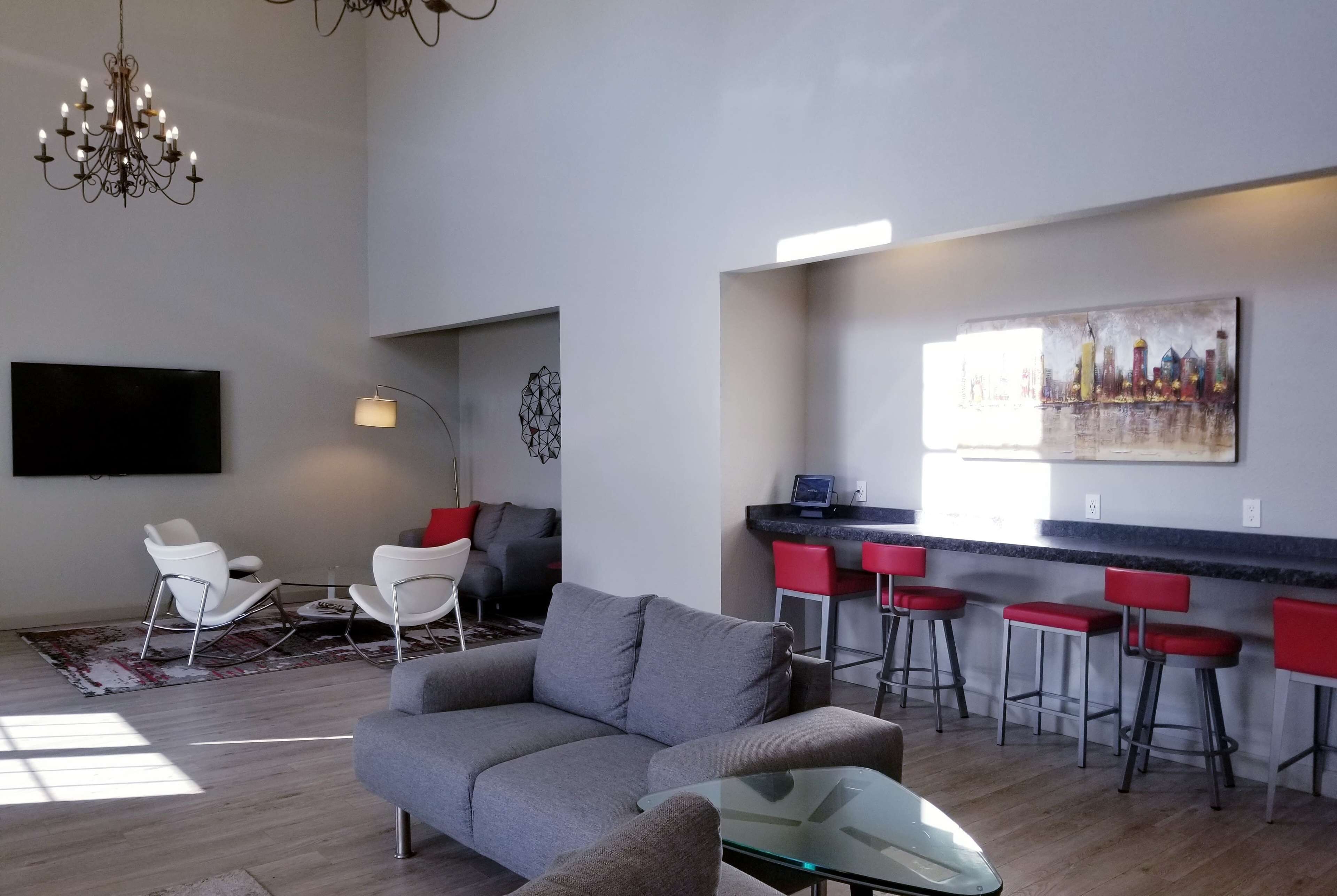 A modern living room with a grey sofa, white chairs, a glass coffee table, a chandelier, a flat-screen TV, a painting, and a kitchen area with red bar stools.