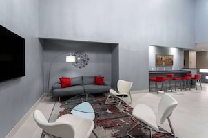 Un salón moderno con un sofá gris, cojines rojos, una mesa de centro de vidrio y sillas blancas. Un televisor de pantalla plana está montado en la pared.