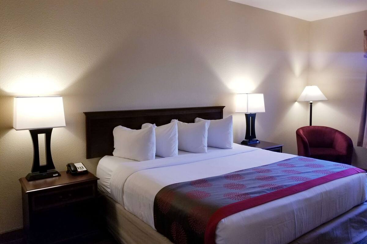 Quarto de hotel com uma cama, duas mesas de cabeceira, uma luminária, uma cadeira e um telefone em uma das mesas.