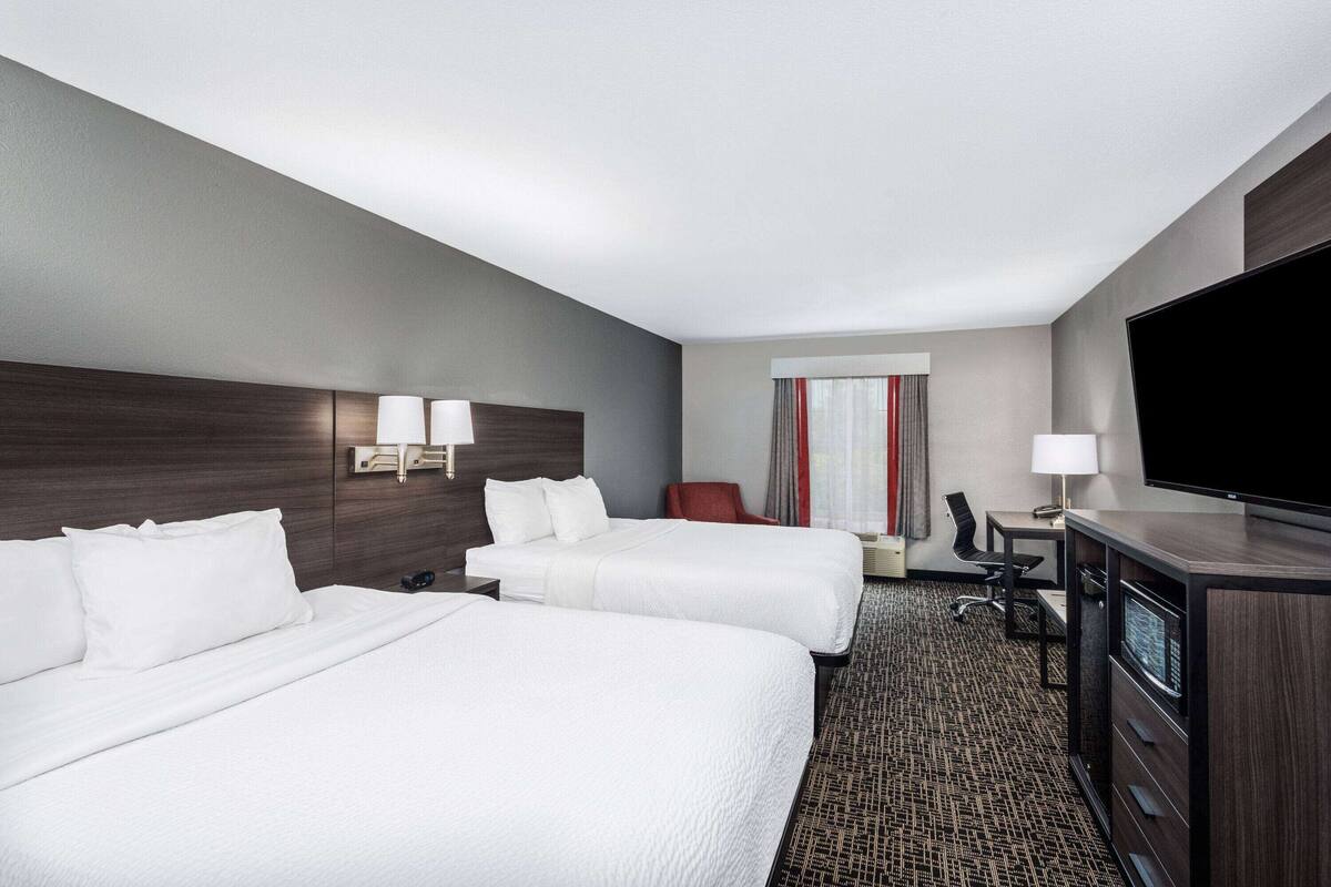 Quarto de hotel com duas camas, uma escrivaninha, uma cadeira, televisão e janela com cortinas.