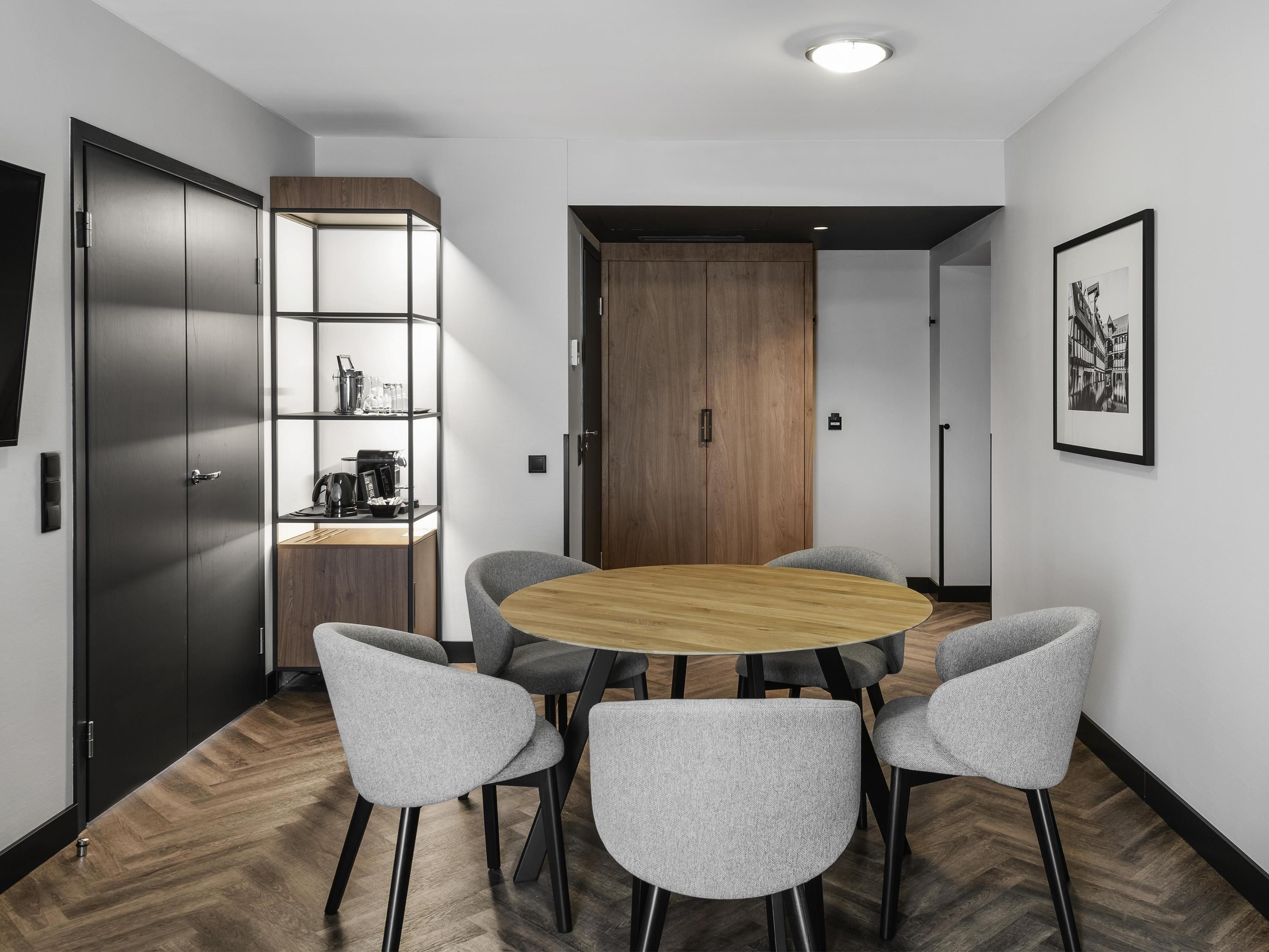 A modern conference room with a round table, grey upholstered chairs, a wooden cabinet with shelves, and a framed picture on the wall.