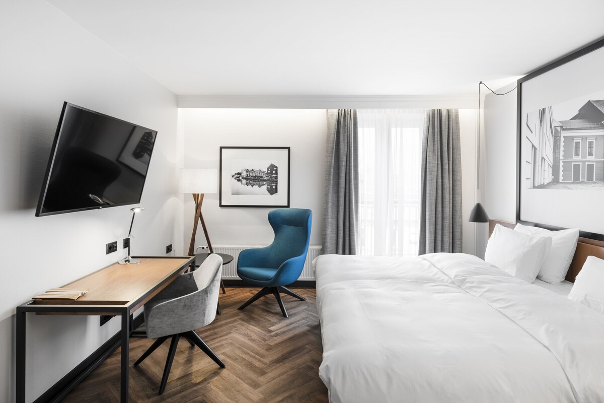 A hotel room with a bed, a desk, a chair, a TV, and a framed picture on the wall.