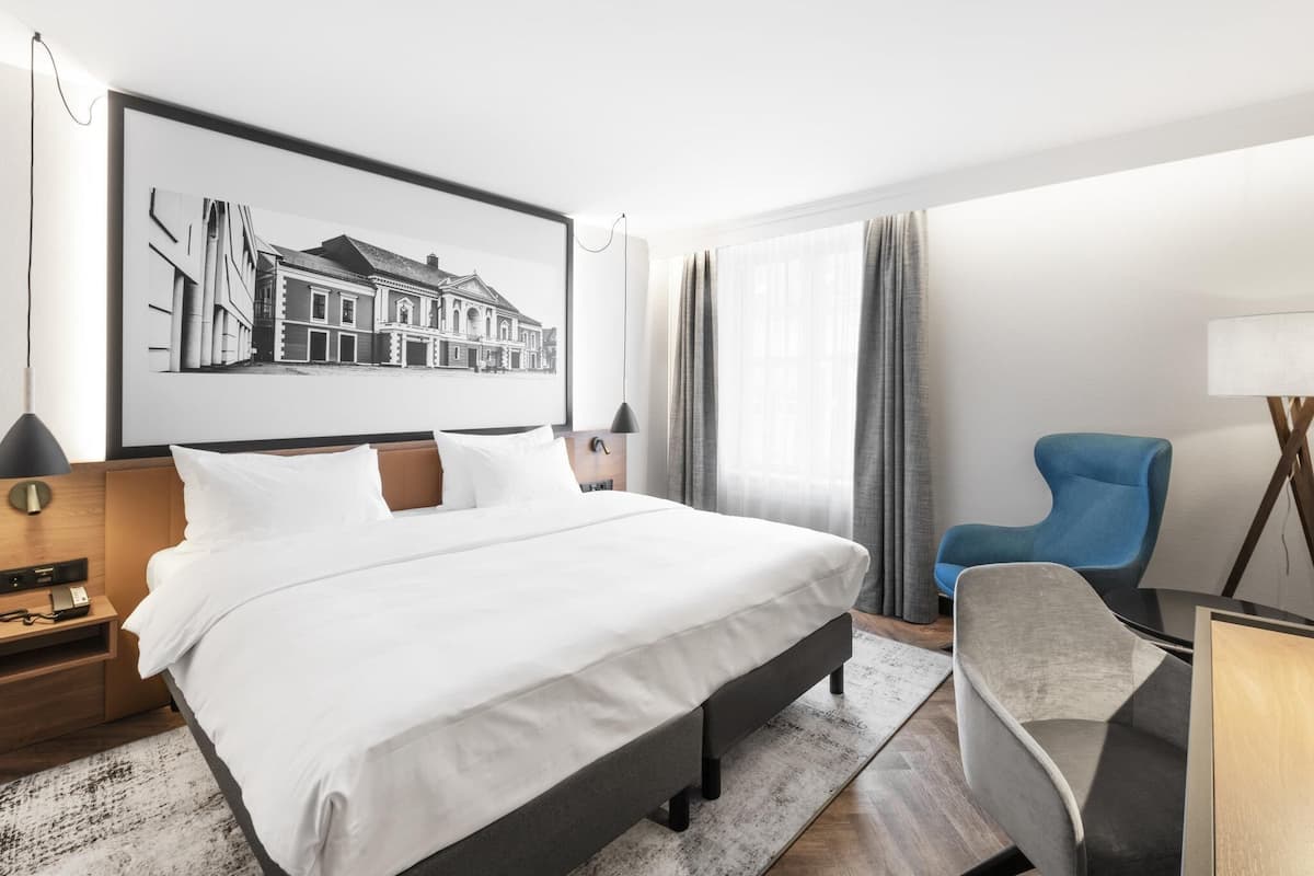 Quarto de hotel com uma cama grande, uma escrivaninha, uma cadeira, um abajur e um quadro na parede.
