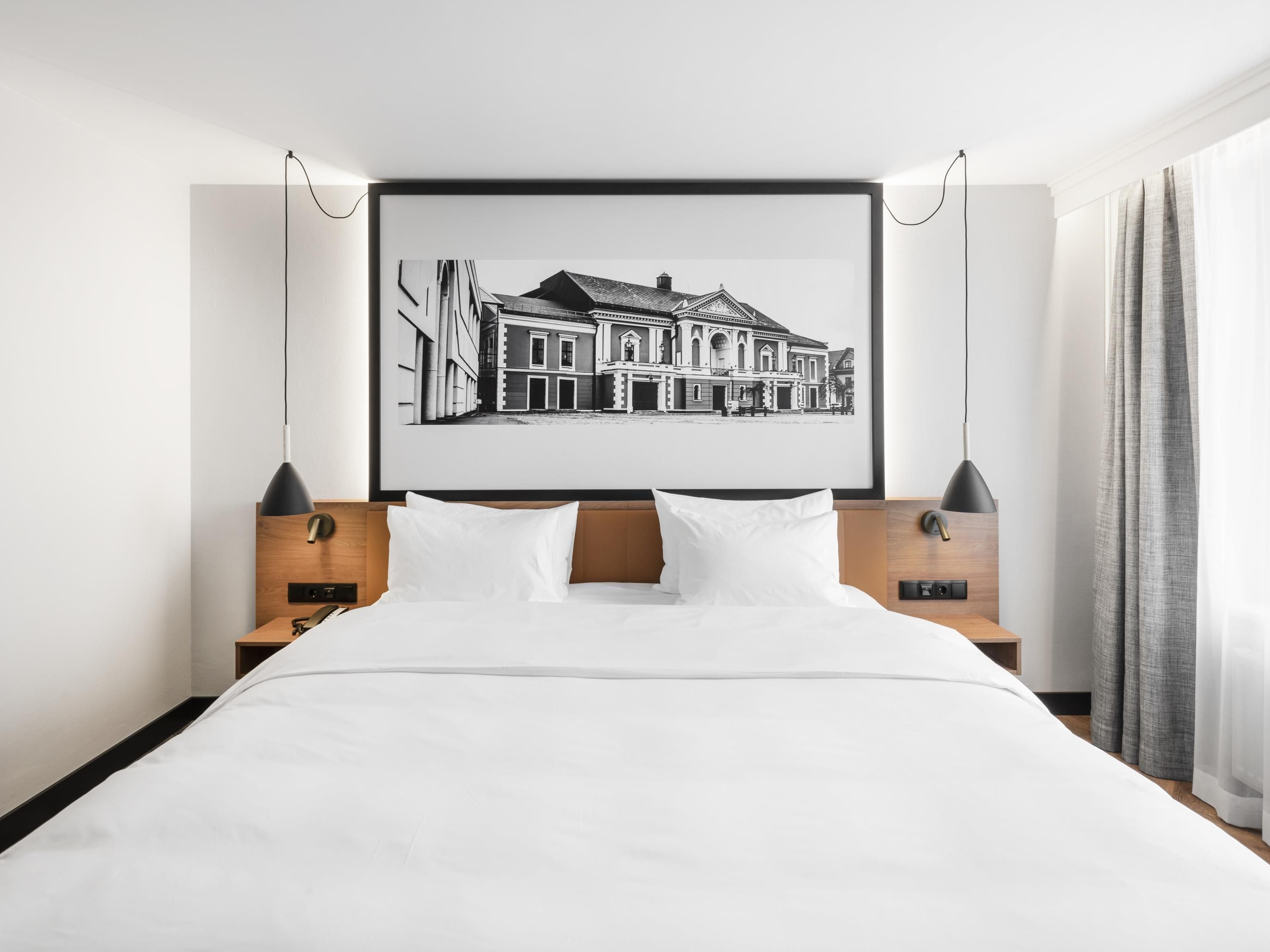 A modern hotel room with a large bed, a framed black and white cityscape artwork, wooden headboard, and two bedside lamps.