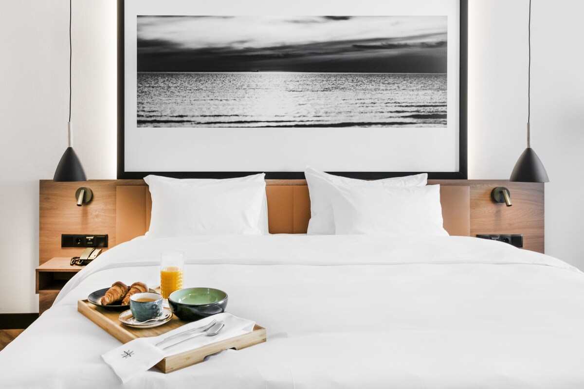 A neatly made bed with white linens, a breakfast tray with croissants, a bowl, and a glass of orange juice. A framed black and white photograph of a beach is on the wall behind the bed.