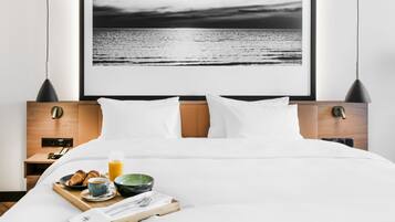 A neatly made bed with white linens, a breakfast tray with croissants, a bowl, and a glass of orange juice. A framed black and white photograph of a beach is on the wall behind the bed.