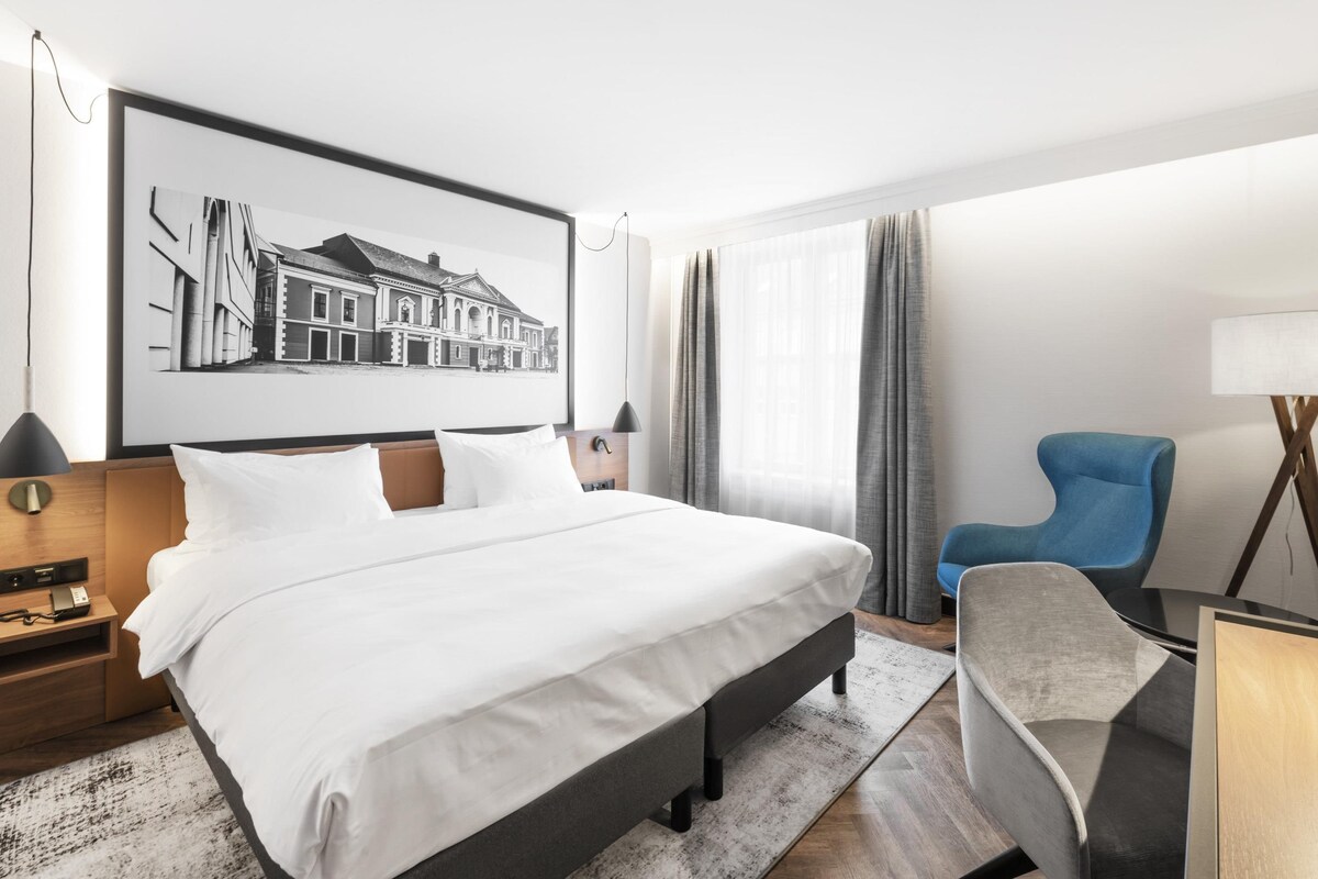 Una habitación de hotel moderna con una cama grande, un sillón azul, un escritorio con lámpara y un cuadro enmarcado en la pared.