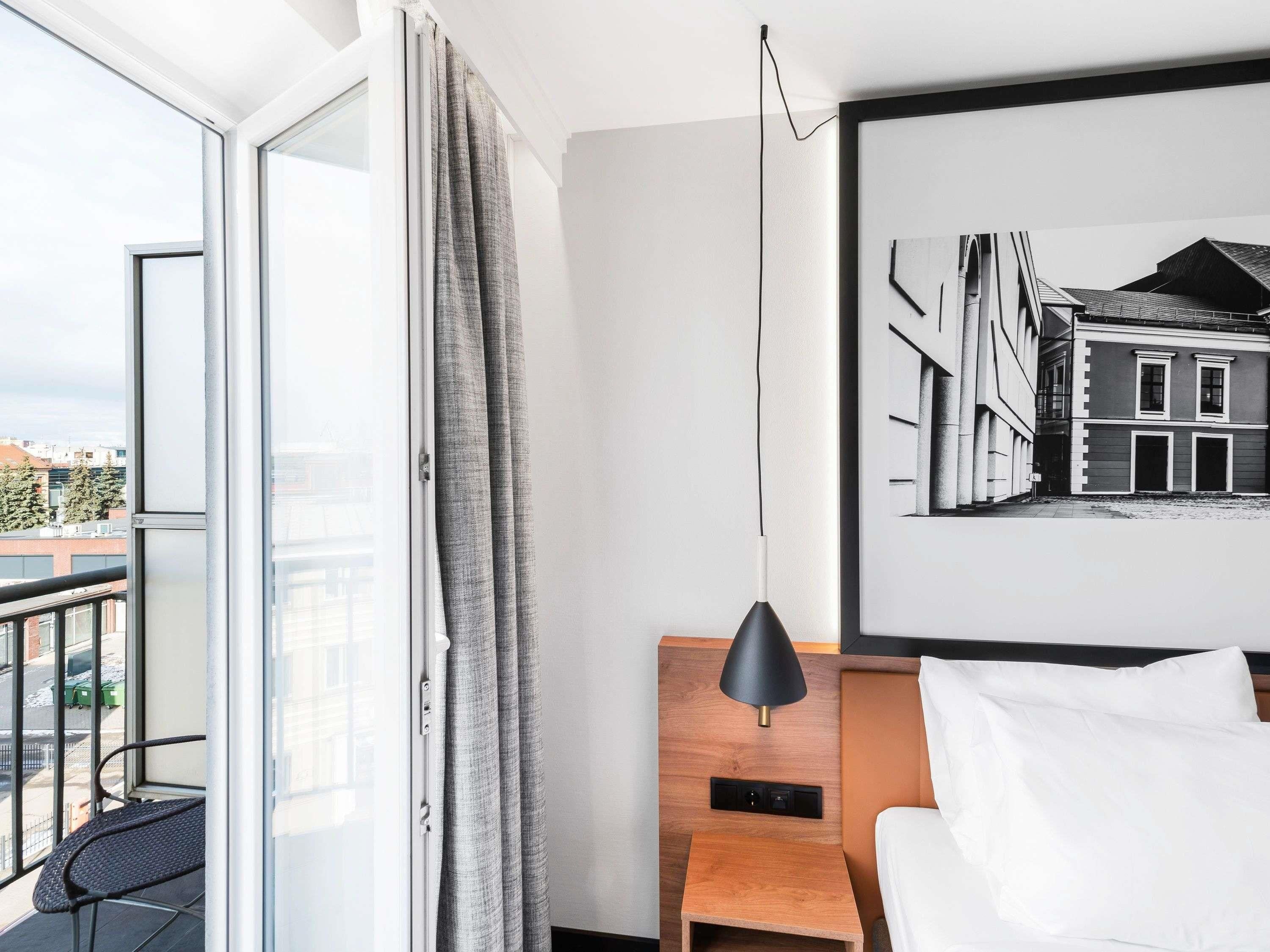 A hotel room with a large window, a framed black and white photograph on the wall, a bedside table with a lamp, and a bed with white bedding.