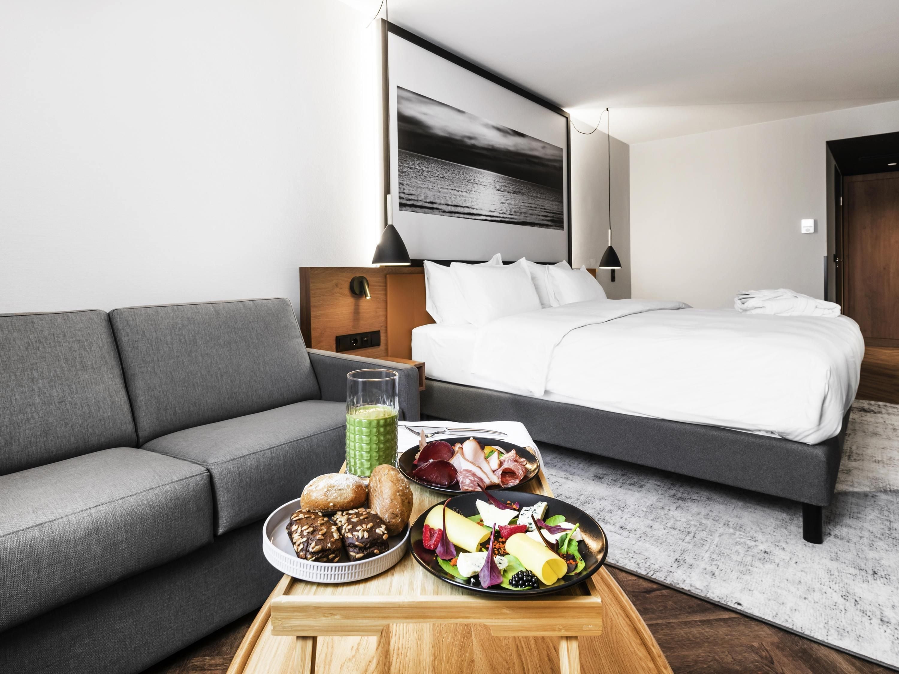 A modern hotel room with a grey sofa, a bed with white bedding, a dining table with food, and a large framed picture on the wall.