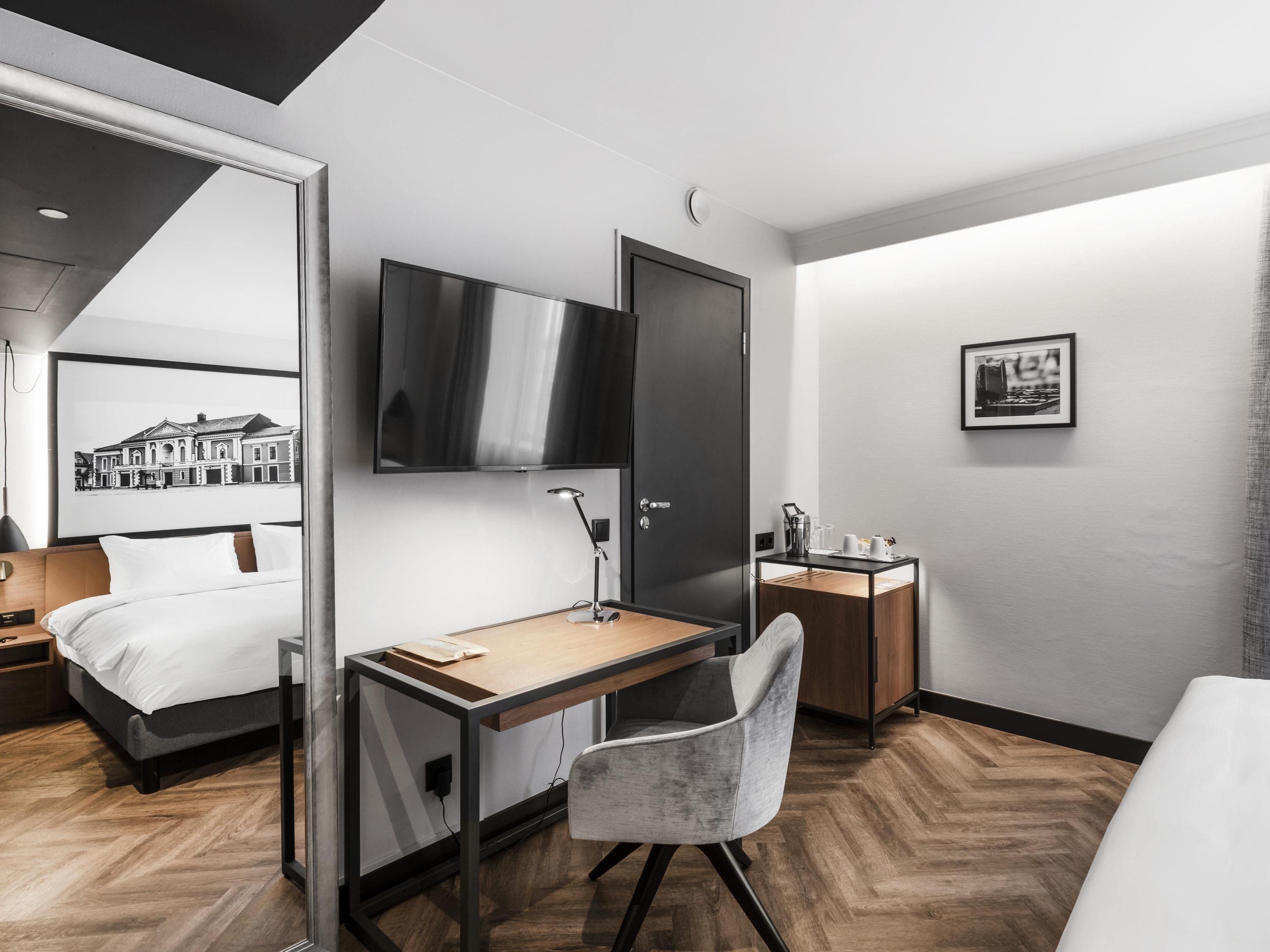 A modern hotel room with a bed, desk, chair, TV, and a small bedside table.