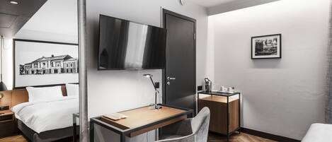 A modern hotel room with a bed, desk, chair, TV, and a small bedside table.
