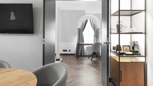 A modern room with a wooden dining table, a grey upholstered chair, a television mounted on the wall, and a built-in coffee station with a coffee maker and cups. - Mercure Klaipeda City (Klaipeda)