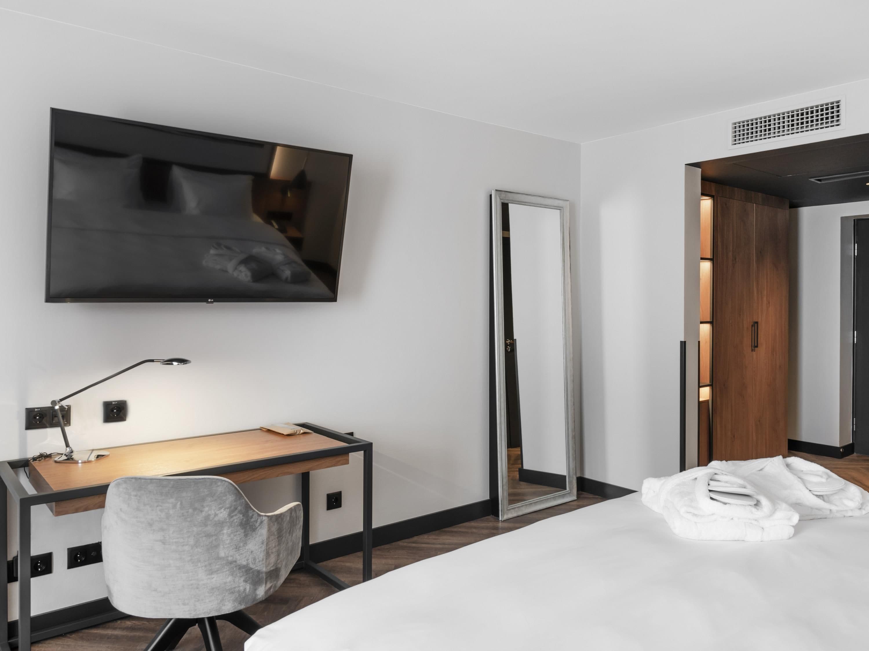A modern hotel room with a large bed, a desk with a chair, a flat-screen TV mounted on the wall, a full-length mirror, and a wooden wardrobe.