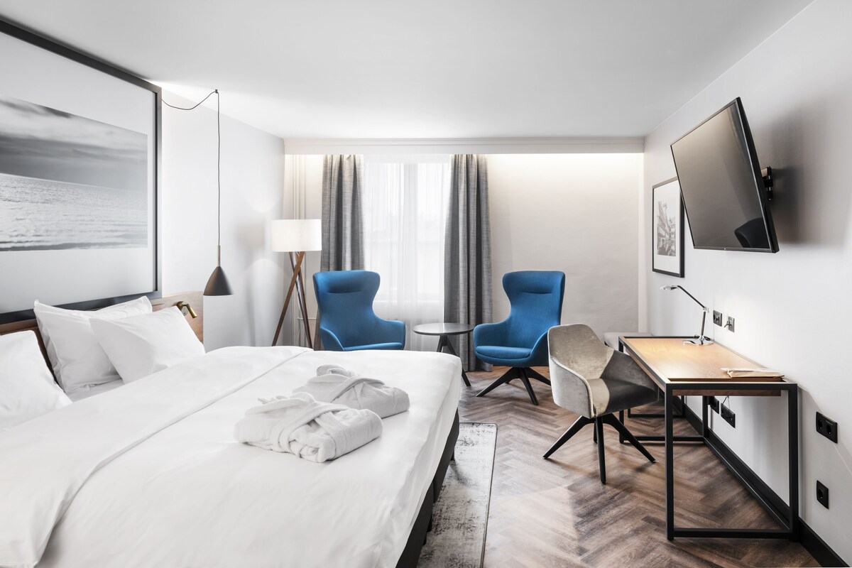 A hotel room with a bed, two blue chairs, a desk, a TV, and a large window with curtains.