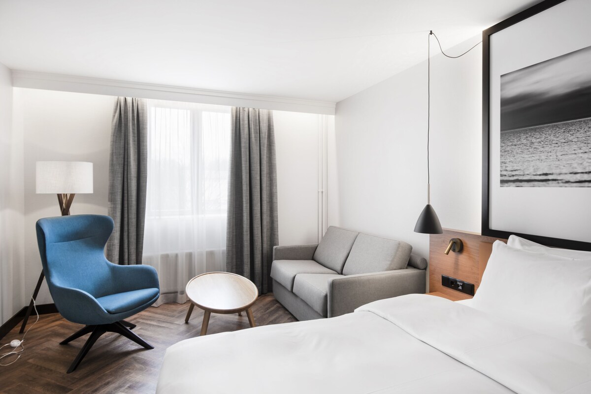 A hotel room with a bed, a grey sofa, a blue armchair, a small round table, and a window with curtains.