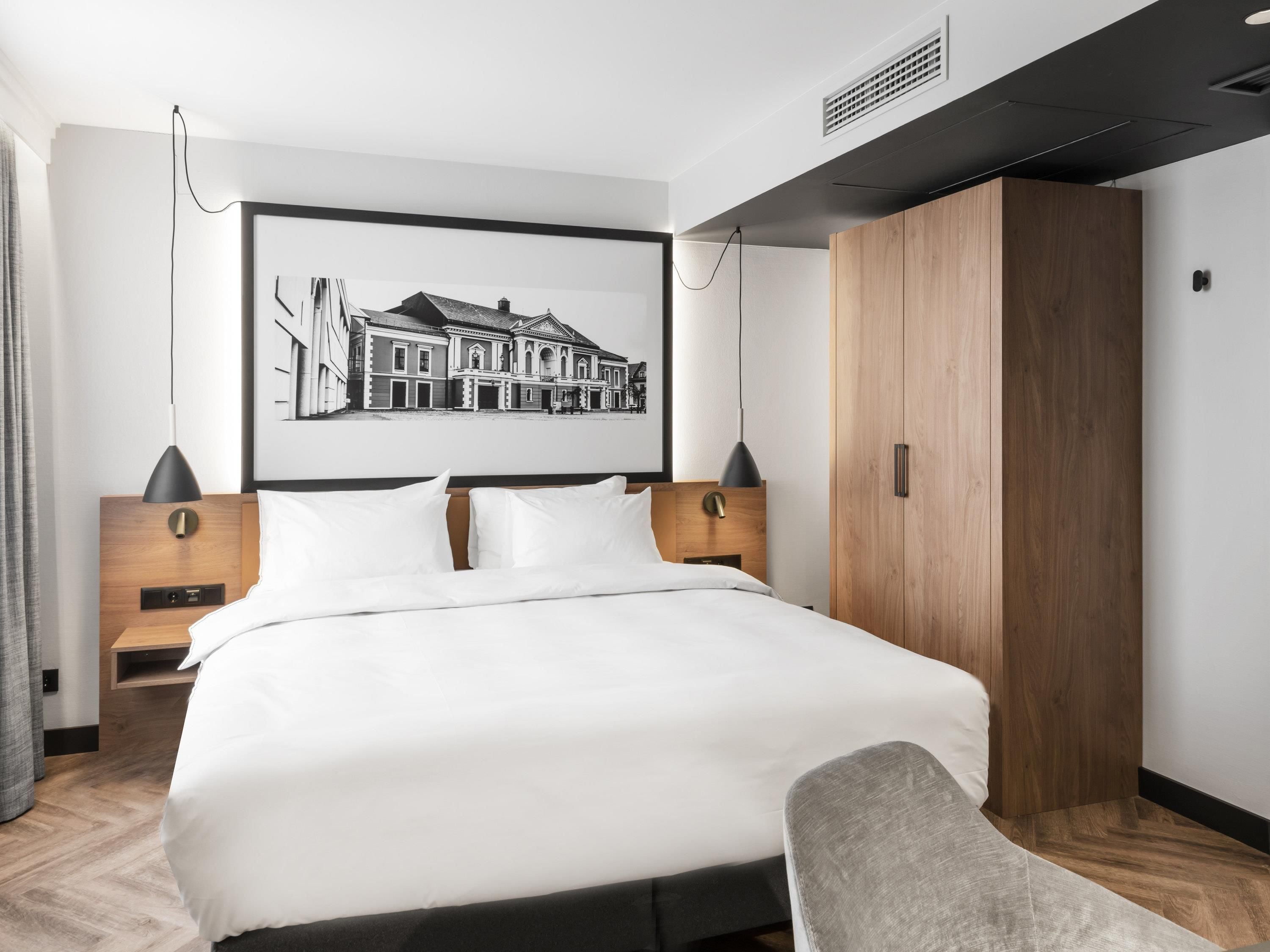 A modern hotel room with a large bed, wooden wardrobe, and a framed cityscape picture on the wall.