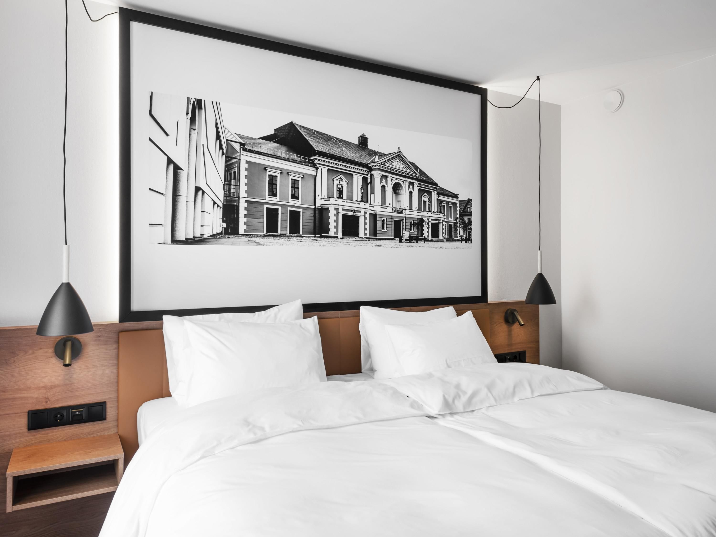Une chambre d’hôtel moderne avec une grande photographie en noir et blanc, encadrée, représentant un bâtiment historique, un lit soigneusement fait et deux lampes de chevet.