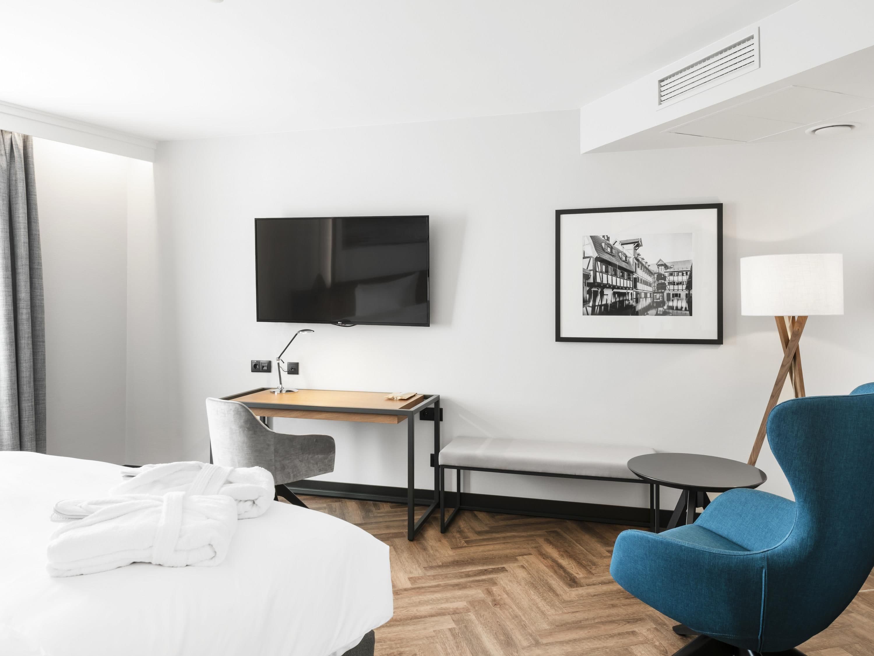 A hotel room with a bed, a desk with a chair, a television, a framed picture, a lamp, and a blue armchair.