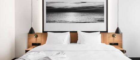 Una habitación de hotel moderna con una cama grande, ropa de cama blanca, dos mesitas de noche con lámparas y una fotografía enmarcada de una playa en la pared.