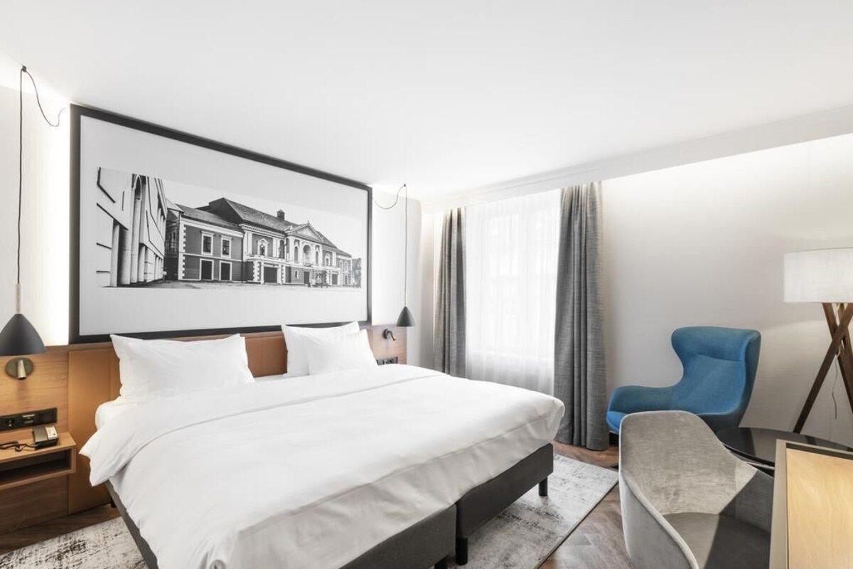 A modern hotel room with a large bed, a desk, a chair, and a framed picture on the wall.