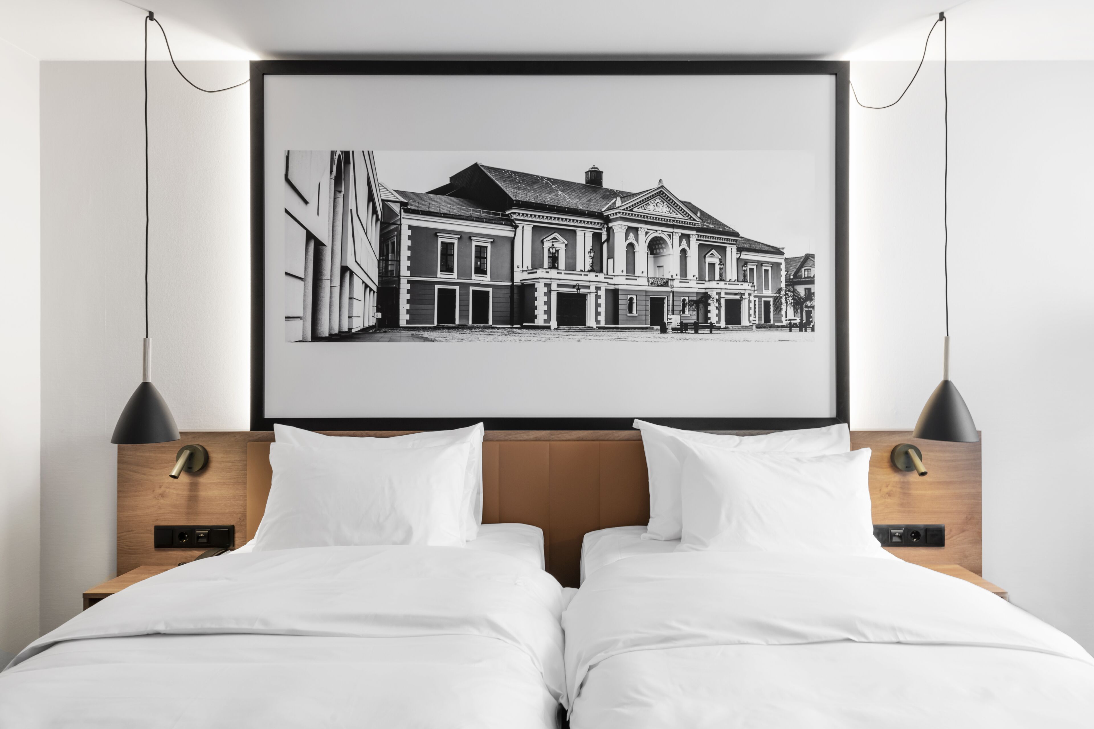 A hotel room with a large bed, two bedside tables, a framed black and white cityscape artwork, and a modern pendant light.