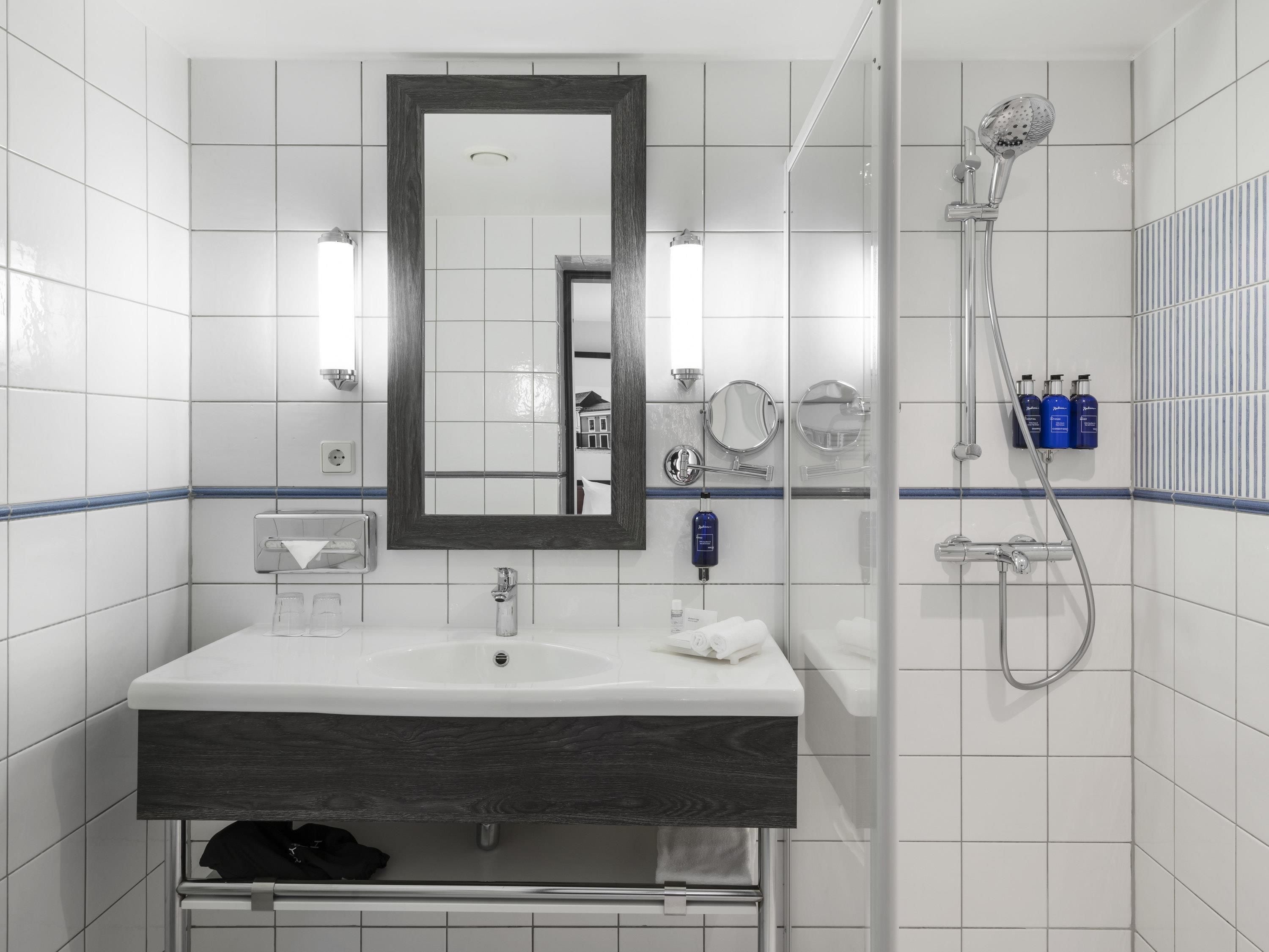 A modern bathroom with a white sink, dark wood vanity, a large mirror, a shower with a handheld showerhead, and toiletries on the wall.