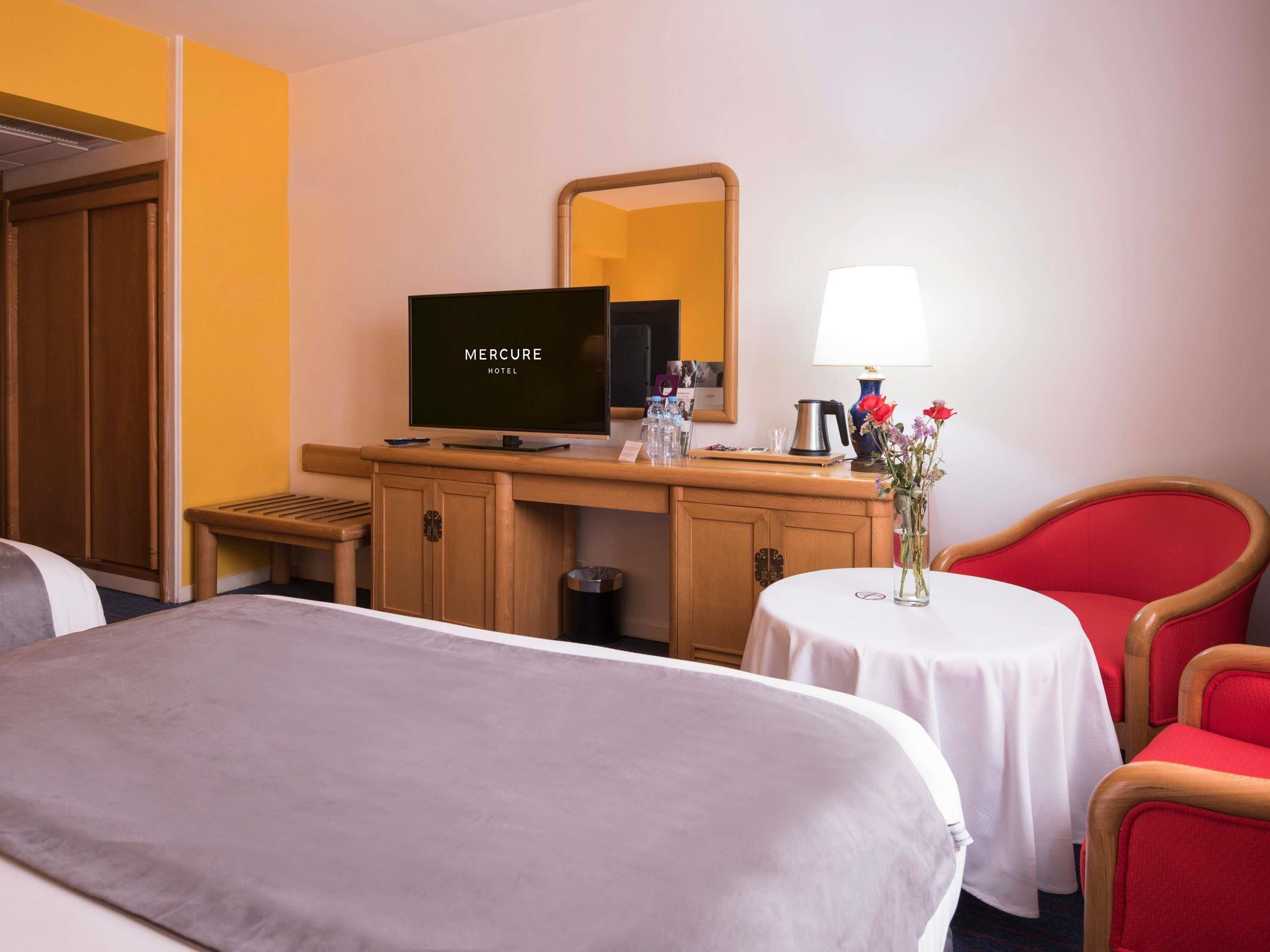 Habitación de hotel con cama, escritorio con televisor mostrando 'MERCURE HOTEL', mesita con lámpara, silla y jarrón con flores.