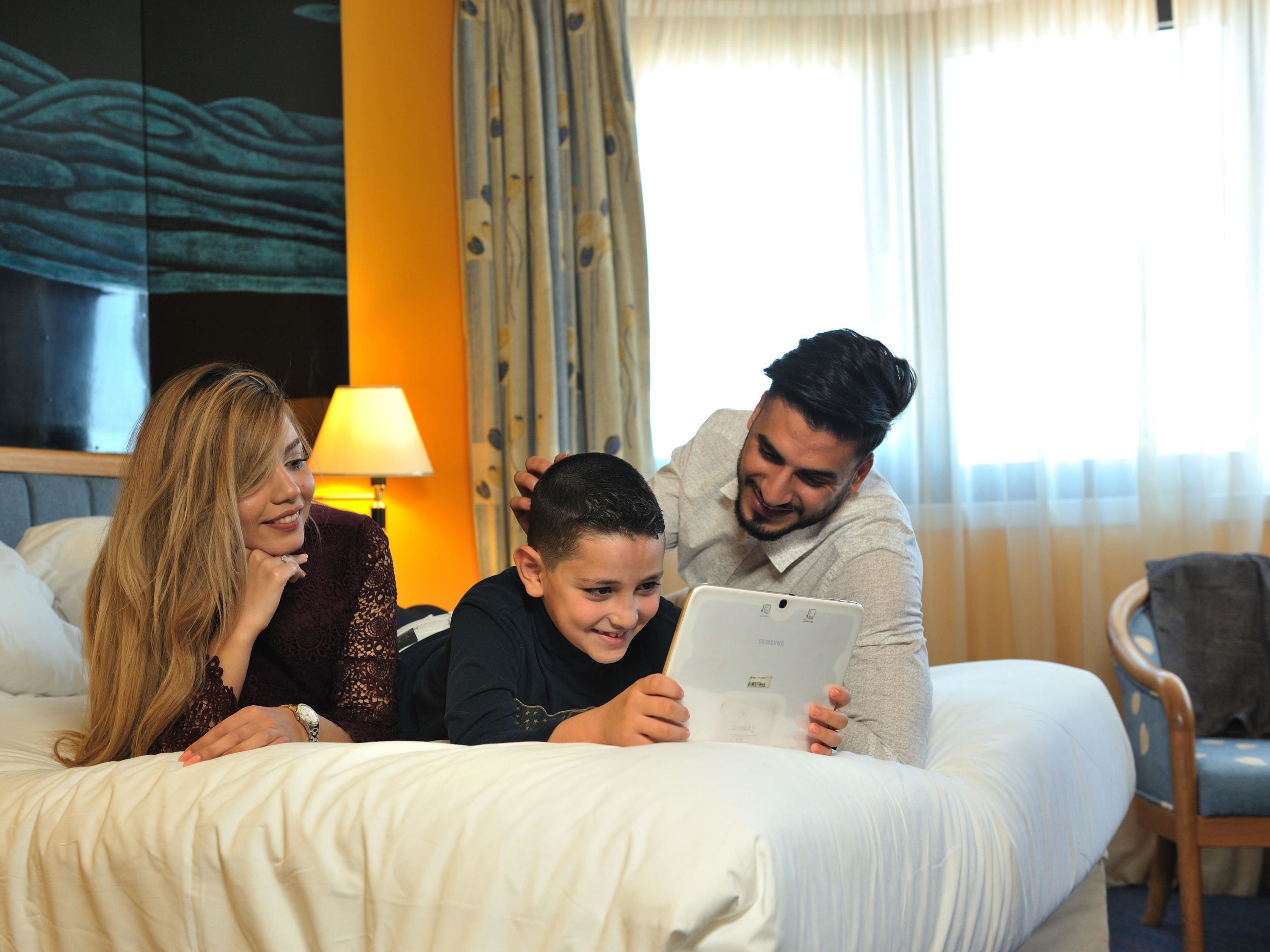 Eine dreiköpfige Familie auf einem Bett, im Hintergrund eine Lampe und Vorhänge.