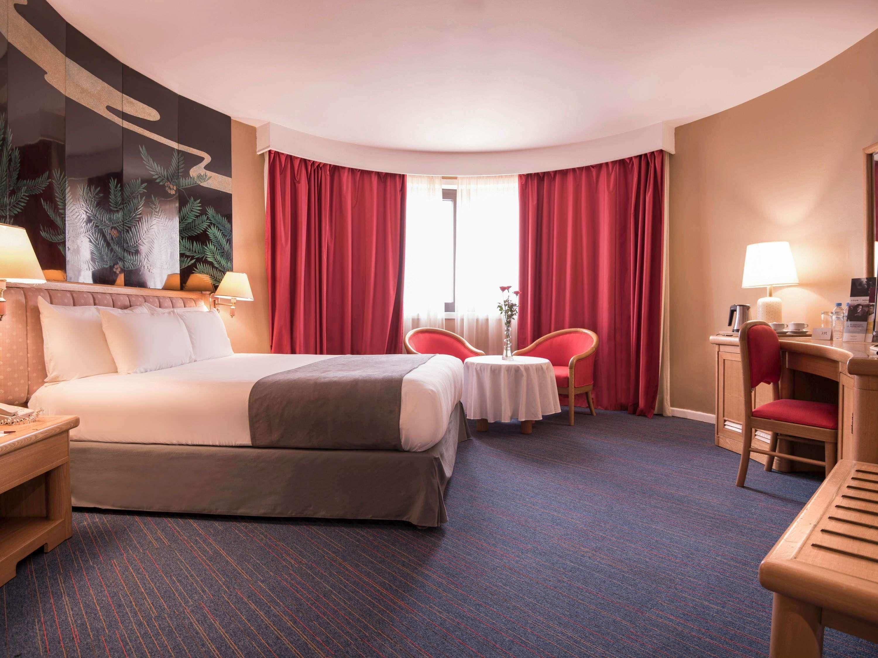 Habitación de hotel con una cama grande, un escritorio, dos sillas, una mesita y un ventanal con cortinas rojas.
