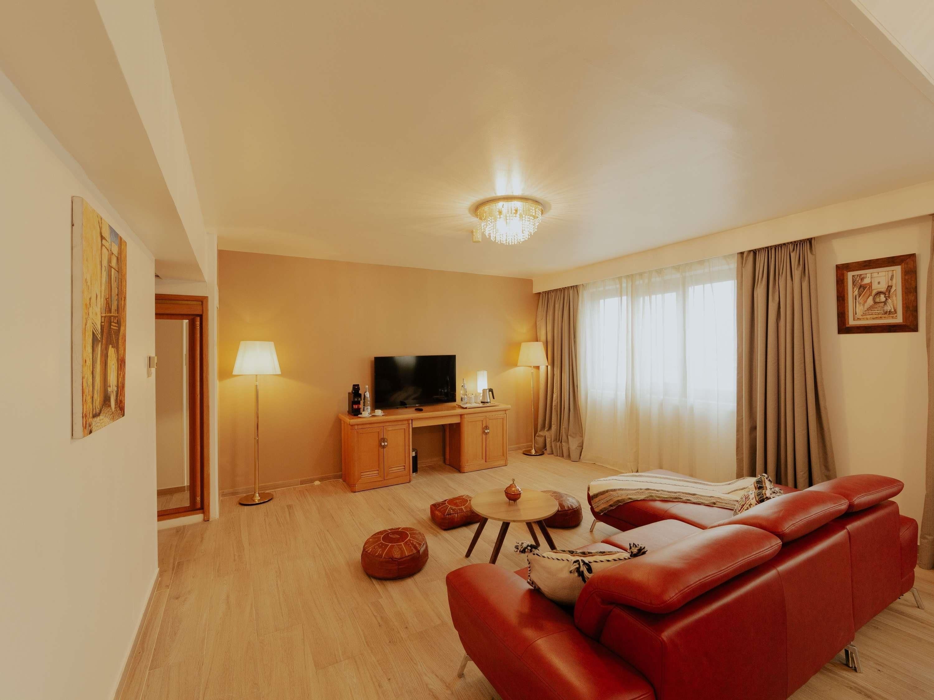 Habitación de hotel con sofá rojo, televisor, armario, lámpara y ventana grande con cortinas.