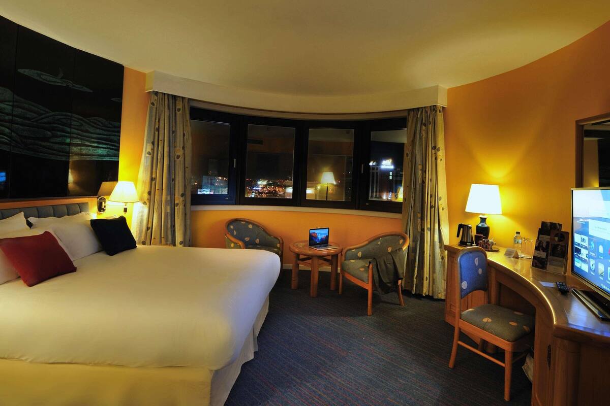 A hotel room with a large bed, a desk with a computer, a chair, and a view of the city at night.