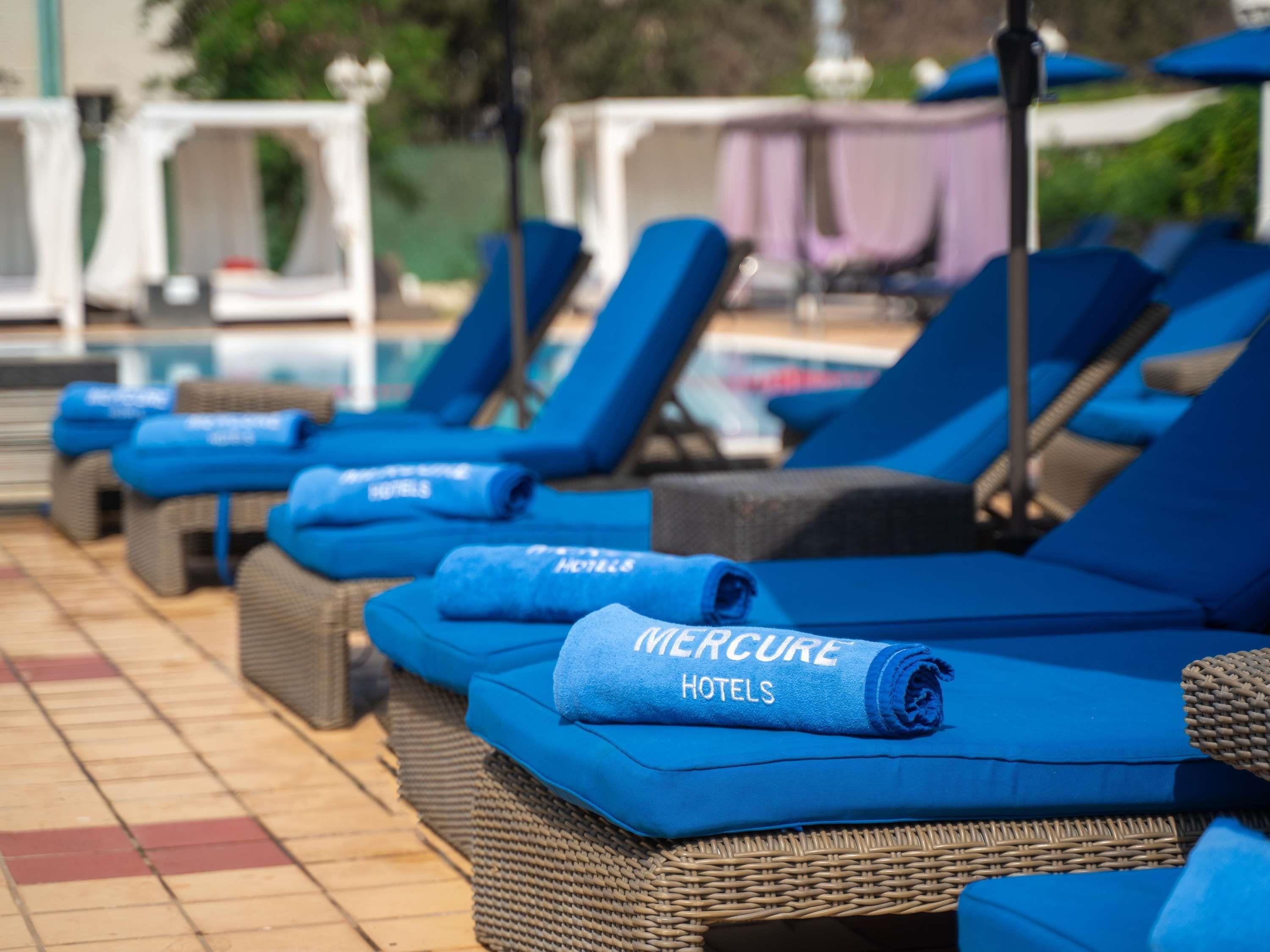 Une rangée de chaises longues avec des coussins bleus, et une serviette sur laquelle on peut lire « Hôtels Mercure ».