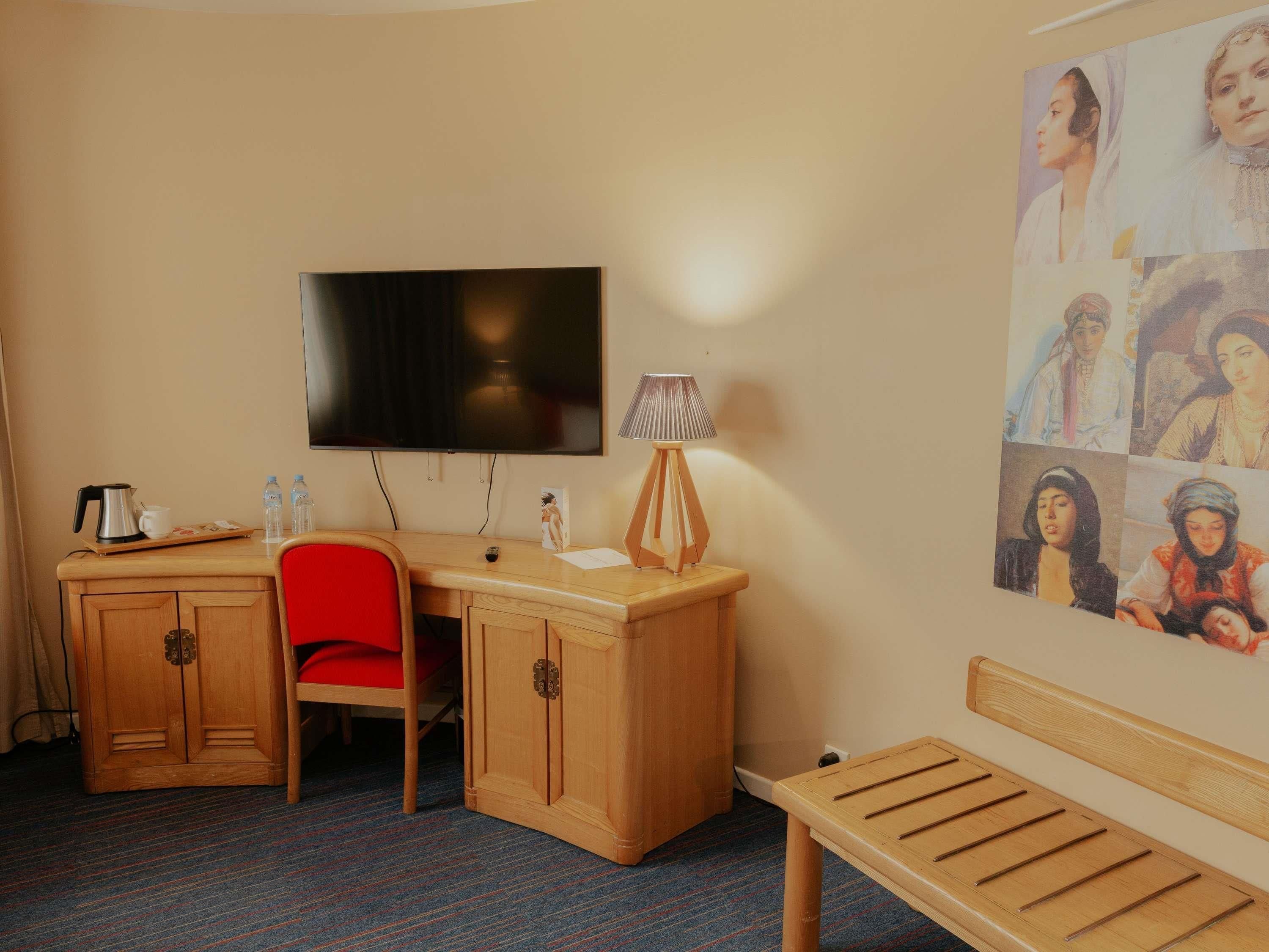 Habitación de hotel con un escritorio de madera, una silla roja, un televisor, una lámpara, un banco y una pared con un collage de imágenes.