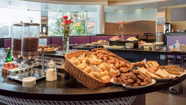 A buffet counter with various food items, a coffee machine, and a decorative vase with flowers. - Mercure Alger Aeroport (Algiers)
