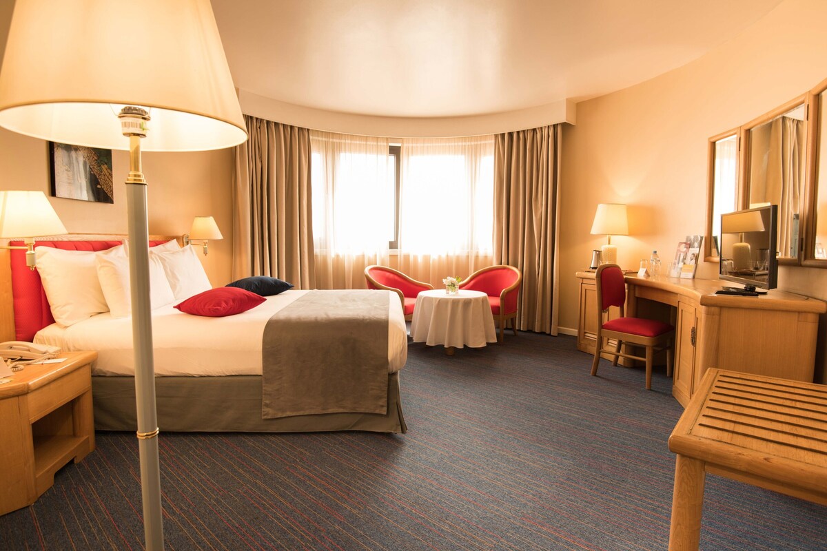 A hotel room with a bed, bedside tables, a desk, a chair, a small table with a vase, and a television.