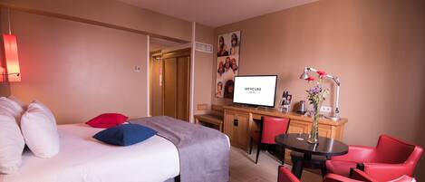 Una habitación de hotel con una cama, un escritorio, un televisor, una silla y una pequeña mesa con un florero.