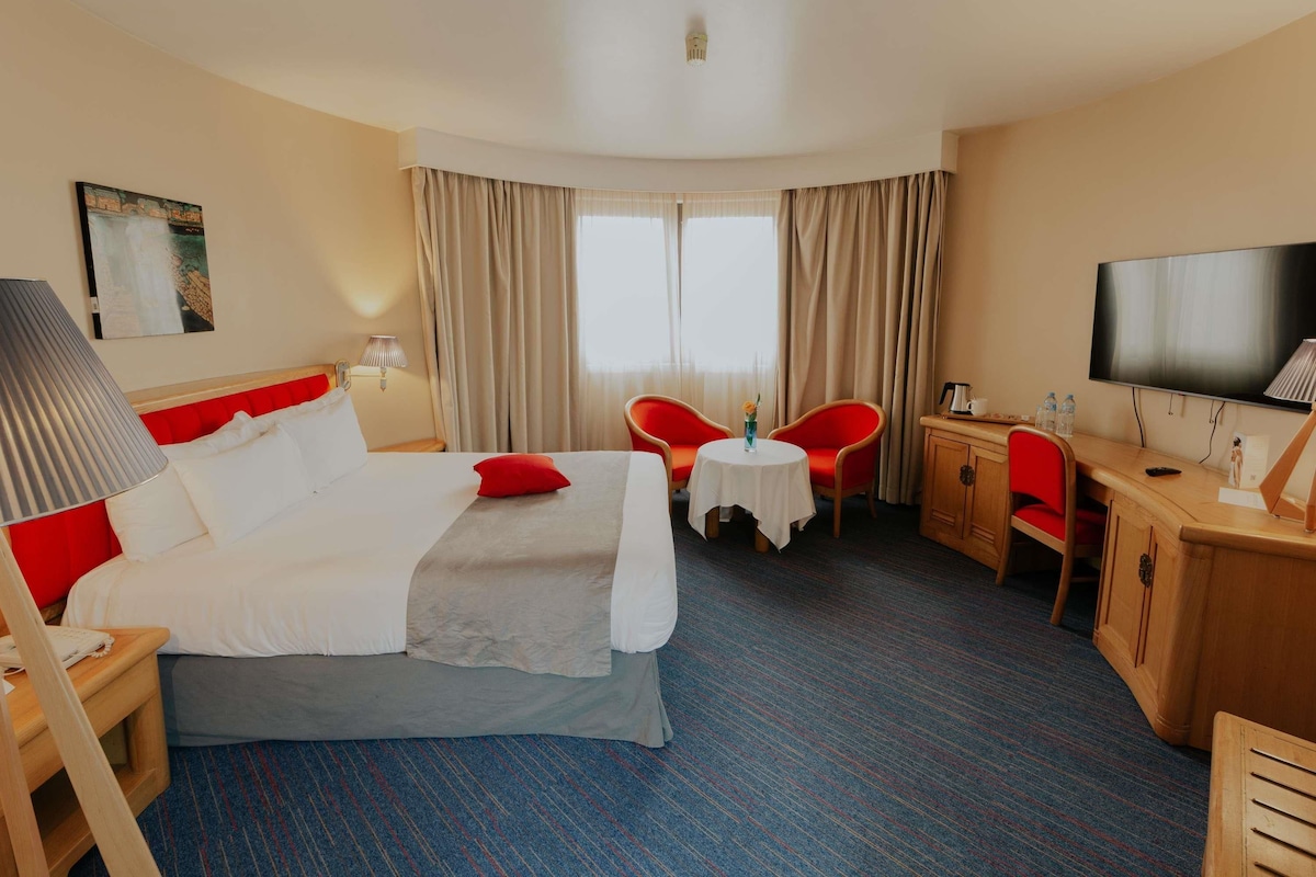 Habitación de hotel con cama, escritorio, silla, televisor y ventana con cortinas.