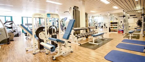 Un gimnasio con diferentes equipos de ejercicio, pisos de madera y colchonetas azules.