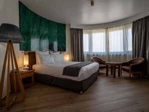 Une chambre d’hôtel avec un lit, une lampe de chevet, une chaise, une table et une grande tapisserie verte.